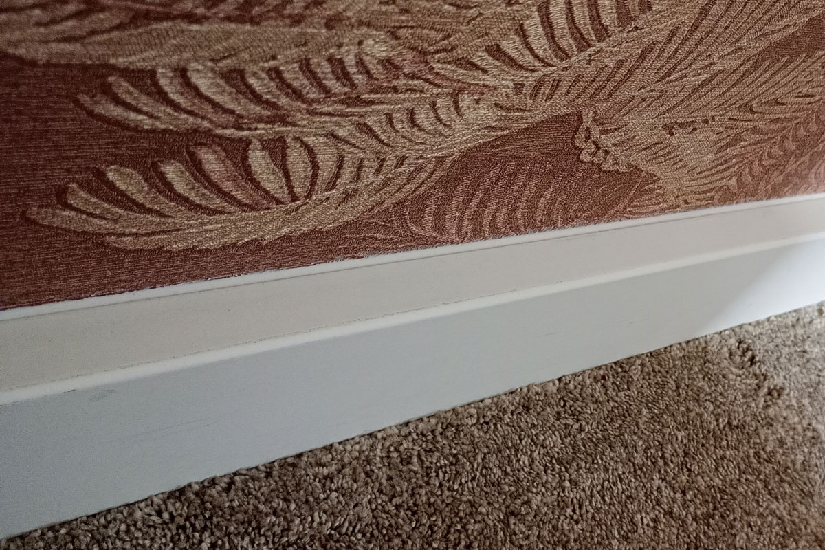 Plinten kitten: witte plint met kitnaad tegen een behangen wand, boven vloerbedekking